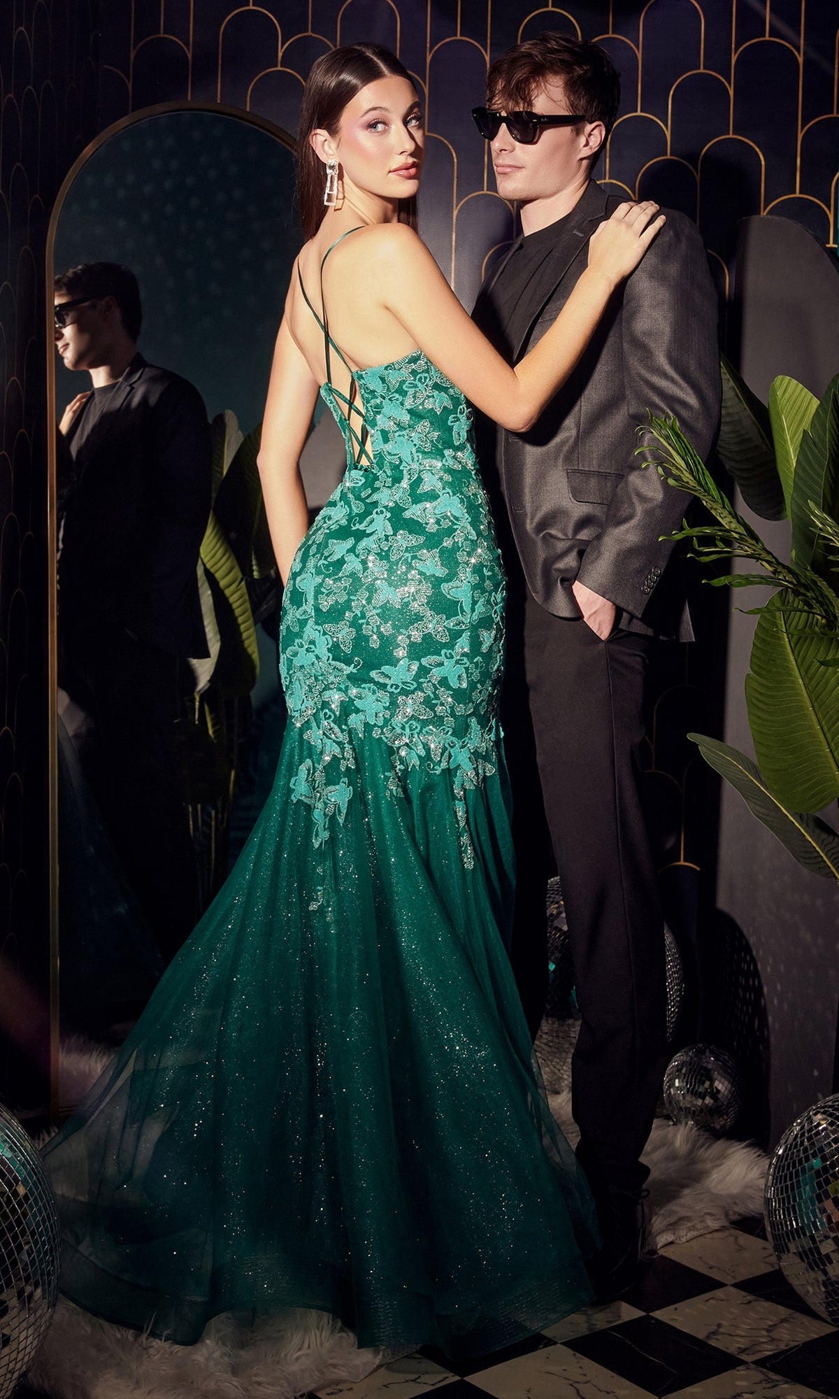 A woman in a Butterfly Emerald Green Mermaid Prom Dress CB112 stands beside a man in a gray suit and sunglasses, with plants and a mirror behind them.